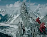 Dwóch alpinistów w czerwonych kurtkach odpoczywa na skalistej grani, podziwiając rozległy widok na lodowiec i ośnieżone szczyty. Broad peak góra w oddali.