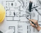 Un arquitecto o proyectista de una obra revisa planos detallados de una casa, con casco amarillo, lápices y calculadora.
