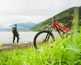 En person med hjälm och ryggsäck står vid en sjö med en mountainbike framför sig. Berg och en stad syns i bakgrunden. Perfekt för att cykla MTB.
