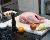 Rohe Gans auf einem Schneidebrett mit Apfel, Zwiebel, Orange, Gewürzen und Pfeffermühle. Die Frage "wie lange dauert auftauen" beschäftigt jeden Koch.