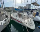 Un voilier First 32, amarré dans un port, avec d'autres bateaux et des bâtiments en arrière-plan.