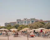 Entspannte Menschen liegen unter Sonnenschirmen am Lido di Venezia Beach. Im Hintergrund thront ein imposantes Gebäude vor üppigem Grün.