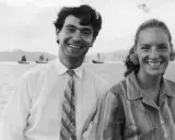 Angela Staude sorride con un uomo su una barca, con velieri sullo sfondo.