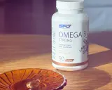 Biała butelka suplementu diety Omega 3 Strong. Na małej, bursztynowej podstawce leży jedna kapsułka. Kwasy omega 3 na co? Na zdrowie!