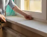 Dwie osoby mierzą taśmą zwymiarową parapet wewnętrzny przy oknie.