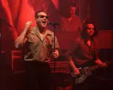 Wokalista w okularach śpiewa na scenie, a basista gra obok. W tle widać klawiszowca. Czerwone światła tworzą atmosferę jak **pale w oknie**.