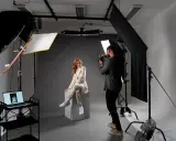 Profesjonalna fotografia studyjna: modelka w jasnym garniturze siedzi na kostce, fotograf robi zdjęcie.