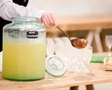 Une louche sert une soupe champenoise sans alcool dans un grand pichet en verre. Des verres à pied et des amuse-bouches sont disposés sur la table.