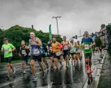 Uczestnicy maratonu biegną w deszczu po mokrej nawierzchni. Widać ich skupienie i determinację mimo niesprzyjającej pogody.