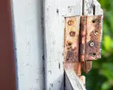 Zardzewiałe zawiasy drzwiowe, które wymagają odrdzewienia śrub. Widać mocno skorodowane metalowe elementy i drewnianą framugę.