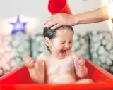 Ein Baby lacht und schreit vor Freude, während seine Haare gewaschen werden. Wasser spritzt, und es hält ein lila Band.