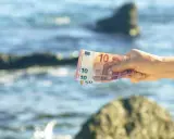 Dłoń trzyma banknoty euro na tle morza. W Czarnogórze obowiązuje euro, więc te pieniądze przydadzą się na wakacjach.