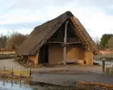 Ein rustikales Bauernhaus aus dem Mittelalter mit Strohdach und Lehmwänden, umgeben von Holzzäunen und Bäumen.