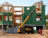 Chantier d'une maison individuelle en cours de construction. Une grue télescopique jaune est positionnée près de la structure en bois et des panneaux verts.