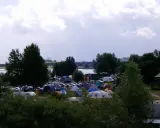 Rozległy kamping z mnóstwem namiotów i samochodów, nad wodą, pod pochmurnym niebem.
