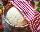 Frischer Pizzateig ruht in einer Schüssel, bedeckt mit einem rot-weiß karierten Tuch. Die Frage: wie viel Hefe in Pizzateig?