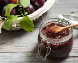 Słoik pełen domowych powideł śliwkowych z czekoladą, obok koszyk z dojrzałymi śliwkami.
