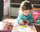 Mała dziewczynka z niepełnosprawnością intelektualną z zaangażowaniem przegląda książeczkę z obrazkami, wskazując paluszkiem na kolorowe ilustracje.