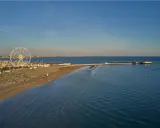 Vista aerea di una spiaggia di Rimini con ruota panoramica, porto turistico e mare calmo sotto un cielo azzurro.
