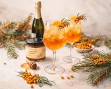 Zwei erfrischende Sanddorn Spritz Cocktails mit Orangenscheiben und Sanddornzweigen, daneben eine Flasche Sekt und ein Glas Sanddorn & Honig.