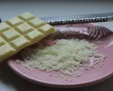 Une râpe à fromage argentée est posée sur une assiette rose, recouverte de copeaux de chocolat blanc fraîchement râpé.