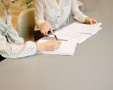 Dwie kobiety omawiają umowę o świadczenie usług. Jedna pisze notatki, druga przegląda dokumenty.