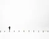 Osoba w czarnym płaszczu balansuje na słupku w śnieżnym krajobrazie. Minimalistyczna, high key fotografia.