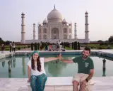 Pareja sonriente en la India, frente al Taj Mahal, con niños y visitantes al fondo.