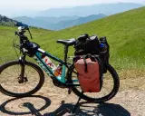Bereit für e bike touren: Ein Trek E-Bike mit Gepäcktaschen steht auf einem Schotterweg, umgeben von grünen Hügeln und Bergketten.