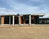 Nowoczesny dom w stanie surowym zamkniętym, z dużymi przeszkleniami i ceglanymi elementami, otoczony placem budowy.