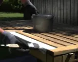 Person med handskar målar en trästol vit. En burk med färg står på bordet.