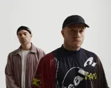 Dwóch mężczyzn, jeden w czapce beanie, drugi w czapce z daszkiem. Na koszulce jednego z nich widoczny jest **guzior fala tekst**.