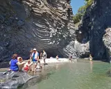 Persone si rilassano e si rinfrescano nelle acque cristalline delle cascate dell'Alcantara, circondate da imponenti pareti rocciose basaltiche.