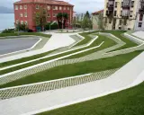 Diseño moderno con césped y caminos de hormigón que forman patrones ondulados. El suelo drenante permite el paso del agua, integrándose con el entorno urbano y el mar al fondo.