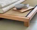 Eine gestapelte, weiße Matratze eines japanischen Bettes ruht auf einer Tatami-Matte. Daneben liegen ein Buch und eine Schale auf einem Holzbrett.