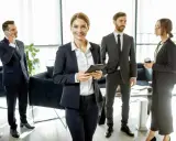 Une femme souriante, possiblement le chief of staff, tient une tablette. Autour d'elle, des collègues en costume discutent dans un bureau moderne.
