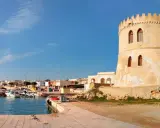 Torre Vado, Lecce: un'antica torre di avvistamento domina un porto con barche e case colorate sotto un cielo azzurro.