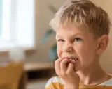 Chłopiec z blond włosami i niebieskimi oczami, który gryzie paznokcie. Może to być objaw stresu lub zespołu Tourette'a.