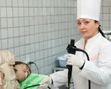 Lekarka w białym fartuchu i czepku przygotowuje pacjenta do gastroskopii, trzymając endoskop.