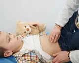 Lekarz bada brzuch dziecka, które skarży się na ból brzucha w okolicy pępka. Obok leży pluszowy miś.
