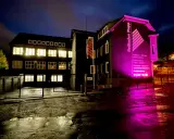 Gebäude mit leuchtenden Fenstern und pinker Beleuchtung. "Rolladen Rapp" wirbt für Nothandkurbel Rolladen, Pflicht für Sicherheit.