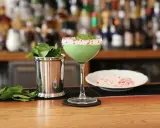 Grön, krämig drink med mynta och krossat strössel på glaskanten. Bredvid står en metallmugg med myntablad och en skål med krossat strössel. Kanske en Amarula drink?