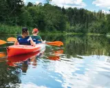 Dwie osoby płyną pomarańczowym kajakiem po spokojnej rzece. Wokół bujna zieleń drzew i trzcin. Idealny spływ kajakowy Mazury.