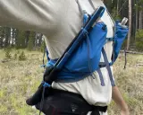 Jak przypiąć kijki do plecaka? Widok z boku na turystę w lesie, z kijami trekkingowymi przymocowanymi do niebieskiego plecaka.