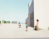 Luigi Ghirri foto: una donna cammina sulla spiaggia, due bambine giocano vicino a un edificio bianco.