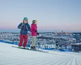 Två skidåkare njuter av utsikten över Hammarbybacken och staden i skymningen.