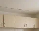Długie pęknięcie na suficie nad jasnymi szafkami kuchennymi. Wygląda, jakby krzywe ściany zaczęły się odsuwać.