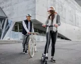 Un homme à vélo et une femme en trottinette, tous deux portant un casque obligatoire, discutent devant un bâtiment moderne.