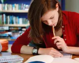 Dziewczyna w czerwonym swetrze myśli nad tekstem rozprawki maturalnej, trzymając długopis przy ustach.