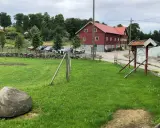 Grönskande gräsmatta med en stor sten framför en röd ladugård på Sammels lantgård. En skylt visar vägen till gårdskafé och butik.